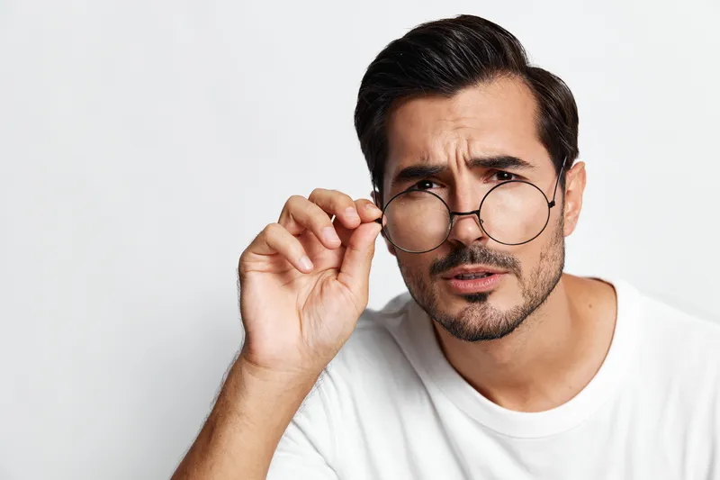 Serious thoughtful young man wearing round glasses, looking at camera with curious expression, isolated on plain solid background. People lifestyle concept