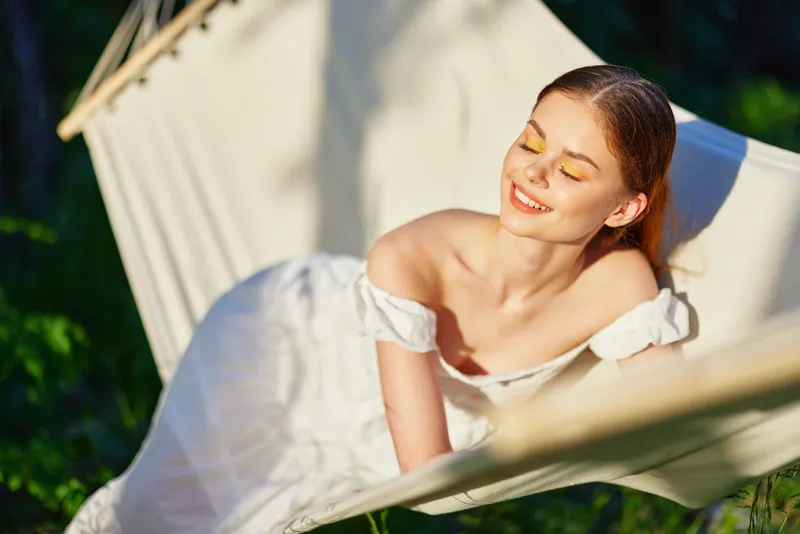 Joyful woman relaxing in a hammock, wearing a white dress and yellow eyeshadow, surrounded by greenery, embodying summer vibes and tranquility