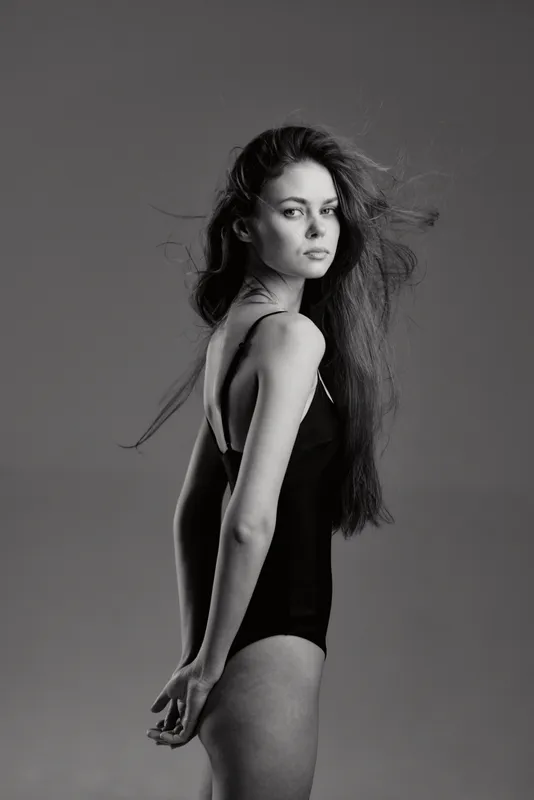 Black and white portrait of a serious woman with long hair in a black bodysuit against a plain background.