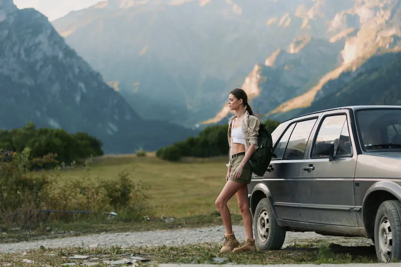 Young woman with backpack standing near car in mountain landscape, casual outfit, enjoying nature, travel adventure, sunny day, outdoor lifestyle, relaxed mood