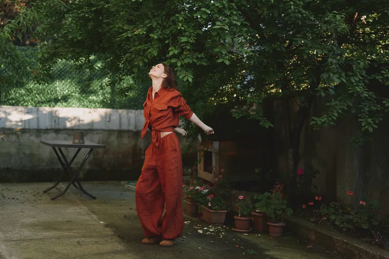 Young woman in a rust red jumpsuit stands in a sunlit garden with arms open, feeling liberated and joyful as greenery frames the scene and a calm courtyard adds serenity