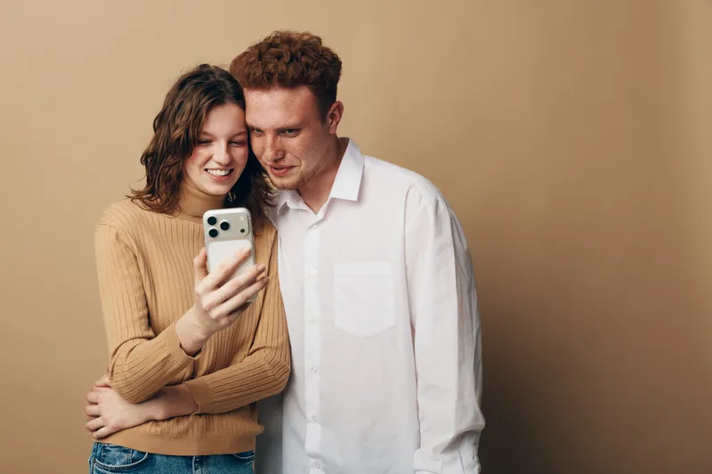 Young couple with casual clothes smiling and looking at smartphone screen together. Warm beige background. Concept of friendship, technology, communication, and happiness.