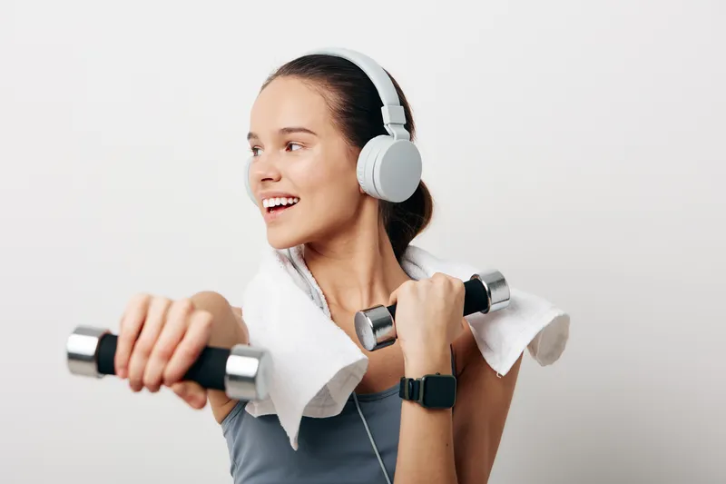 Smiling woman fitness athlete lifting dumbbells with an over the shoulder towel, wearing large headphones and a smartwatch, demonstrating sport training and strength workout indoors on a clean light