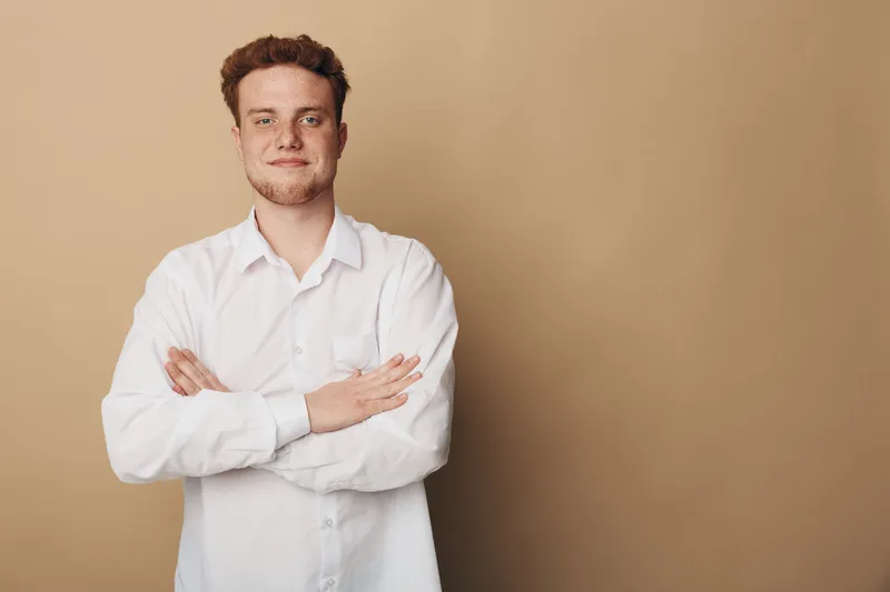 Confident young man with crossed arms wearing white shirt stands against beige background. Casual portrait of smiling male model with short hair and relaxed posture in studio setting.