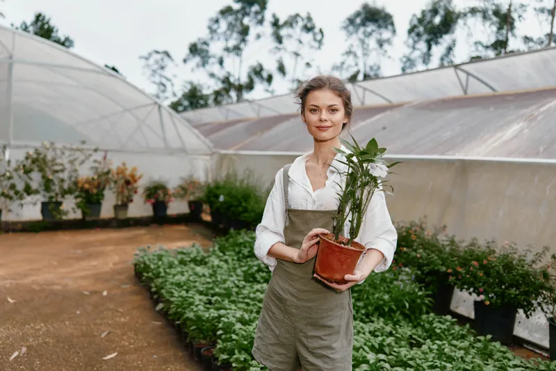 Young woman in a greenhouse holding a potted plant, dressed in a neutral apron, showcasing a connection with nature, happiness, and sustainable gardening