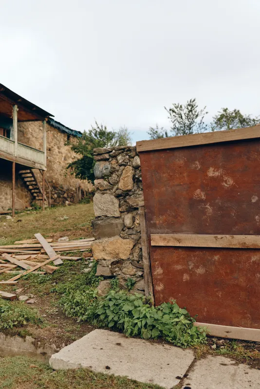 Rural yard with rusted metal gate and stone wall, weathered farmhouse facade, scattered wooden debris and green grass under overcast sky