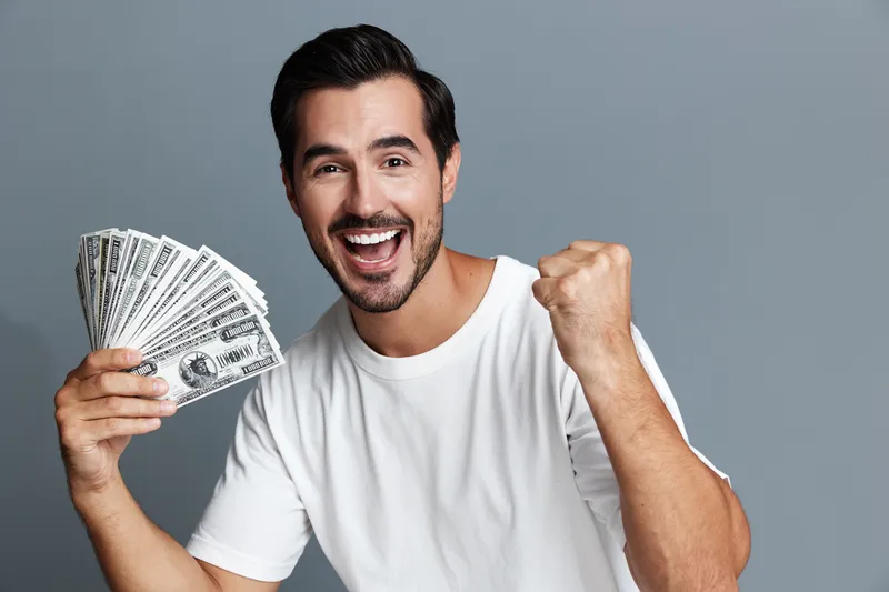 Happy smiling man celebrating with cash money in hand on a solid grey background, expressing joy and excitement. Financial success concept.