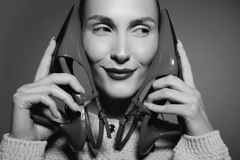 Smiling white woman holds two shoes near her face, wearing sweater. Concept of fashion, beauty, and playful mood in black and white.