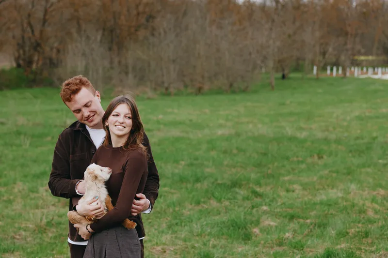 Smiling couple with a fluffy dog embracing in a grassy park, daytime, casual outfits. Love, companionship, and outdoor happiness captured in soft light with a warm, relaxed lifestyle mood.