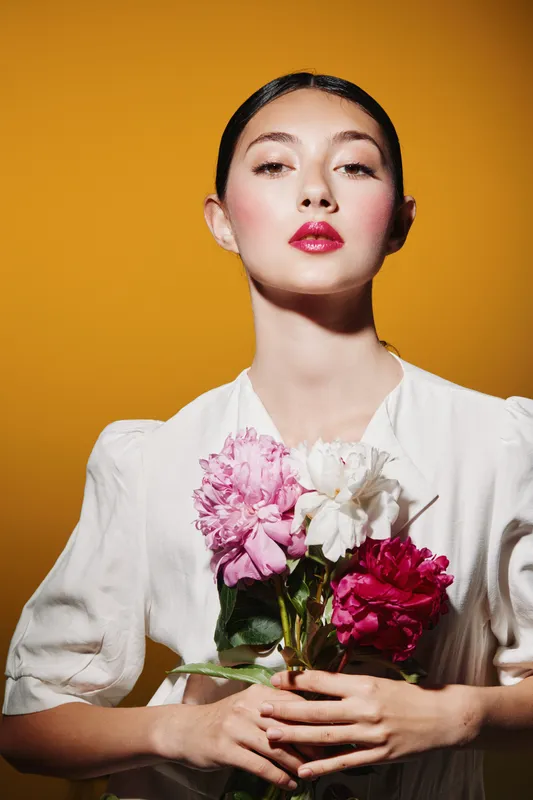Young stylish woman with bold red lipstick poses holding multicolored peonies in white dress on mustard yellow background. Concept of beauty fashion creative flower composition.
