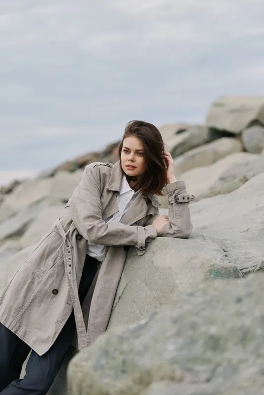 Young woman in a beige trench coat leans on rocks outdoors, looking thoughtful. Overcast sky and natural stone background create a calm, contemplative mood in casual fashion style.