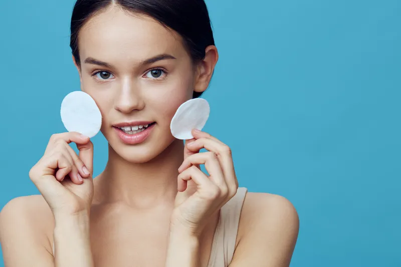 Young woman holding cotton pads, showcasing a fresh skincare routine against a soothing blue background, emphasizing beauty and self care