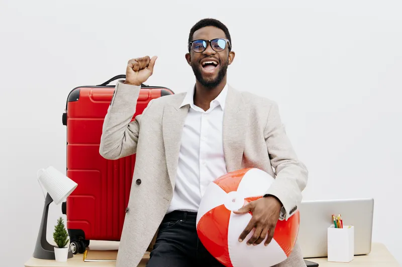 Smiling man with dark skin, sunglasses, and a beige blazer holding a beach ball and standing in front of travel accessories and suitcase, vacation, holiday, summer vibes, travel concept, joyful, leisu