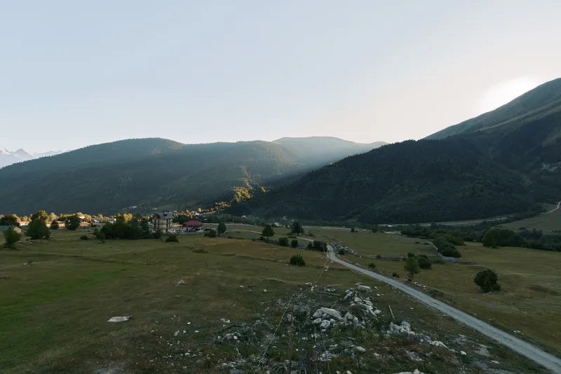 A tranquil mountain valley unfolds with a winding road, gentle fields, and distant hills bathed in warm light. The peaceful scene evokes calm, nature, exploration, and quiet reflection in a wide,