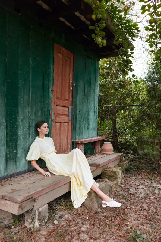 Woman sitting on a wooden porch wearing a pale yellow dress in a rustic outdoor setting, soft sunlight, relaxed pose, tranquil countryside vibe.