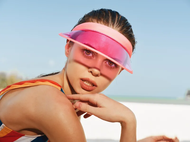 Close-up of a young woman on a sunny beach wearing a pink translucent visor and athletic swimsuit, her confident expression framed by sunlit sea and sand.