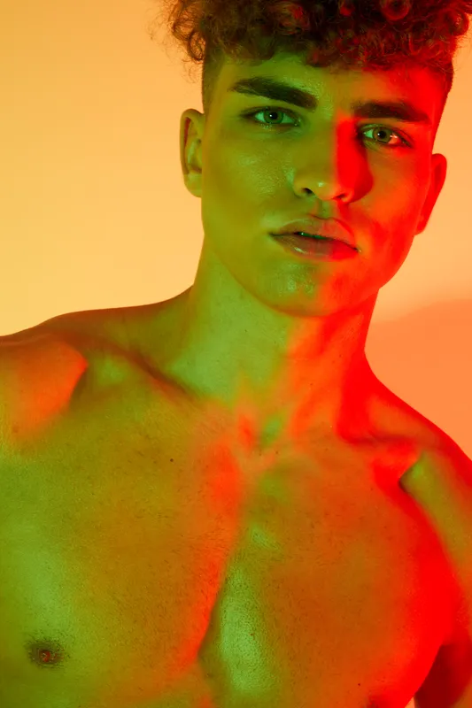 Young man with curly hair and bare chest posing under colorful red and green lighting for artistic portrait photography with dramatic shadows and vibrant tones.