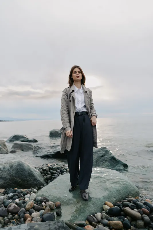 Young woman stands on rocky shore wearing long coat and wide trousers. Overcast sky and calm sea create moody natural outdoor portrait with fashion and nature contrast.
