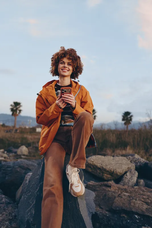Woman lifestyle portrait outdoors, curly hair in orange jacket, smiling and holding smartphone on rocky trail at dusk, casual travel mood, soft natural light