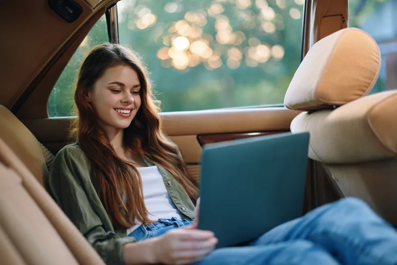Young woman enjoying a laptop in a car with a relaxed expression, wearing casual clothing, and surrounded by a soft bokeh background during sunset.