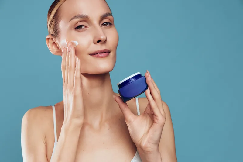 Young woman applying facial moisturizer on her face, holding a skincare jar, calm serene beauty care routine product, studio close up portrait on blue background