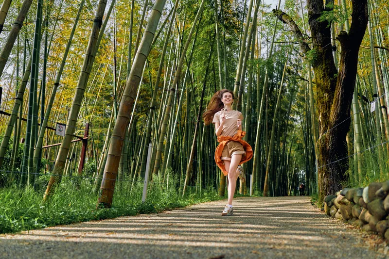 Woman running on a forest path through tall bamboo, smiling in motion wearing orange skirt and casual top. Outdoor nature scene with sunlight, trees and peaceful trail.