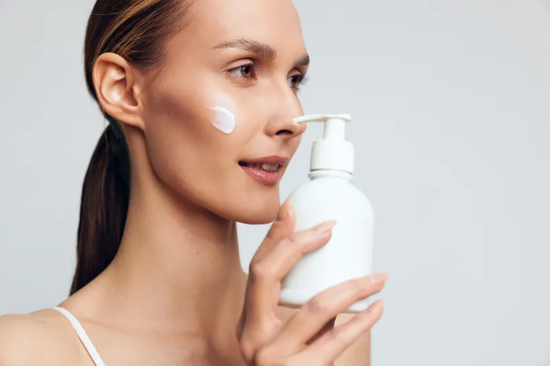 A young woman with clear skin applies white cream on her cheek while holding a white pump bottle. The image represents skincare, beauty routine, and facial moisturizing concepts.