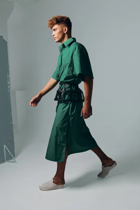 Young man walking in studio wearing green oversized shirt and wide pants with white slip on shoes, side profile and casual fashion style on gray background.