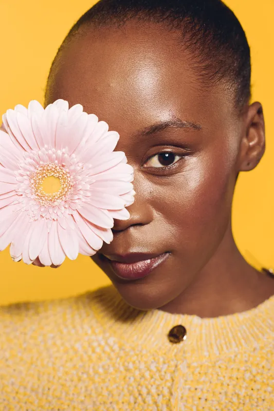 Close-up of a smiling black woman with dark skin holding a pink gerbera flower near her face. Yellow background and yellow sweater create a bright, cheerful mood. Concept of beauty and nature.