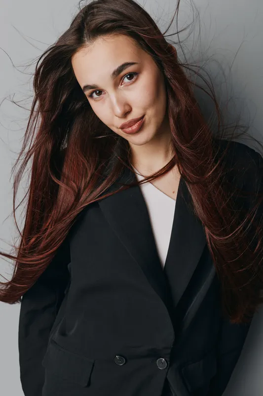 Young woman with long hair wearing black blazer and white shirt posing against gray background. Portrait of confident female model with natural makeup and loose hair in studio lighting.