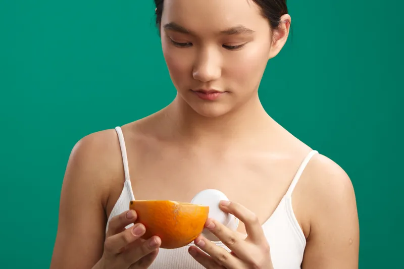asian woman wearing a white top holds an orange half and a white egg, standing against a vivid green backdrop, focused and calm, conveying a healthy, fresh lifestyle in a simple studio setting.