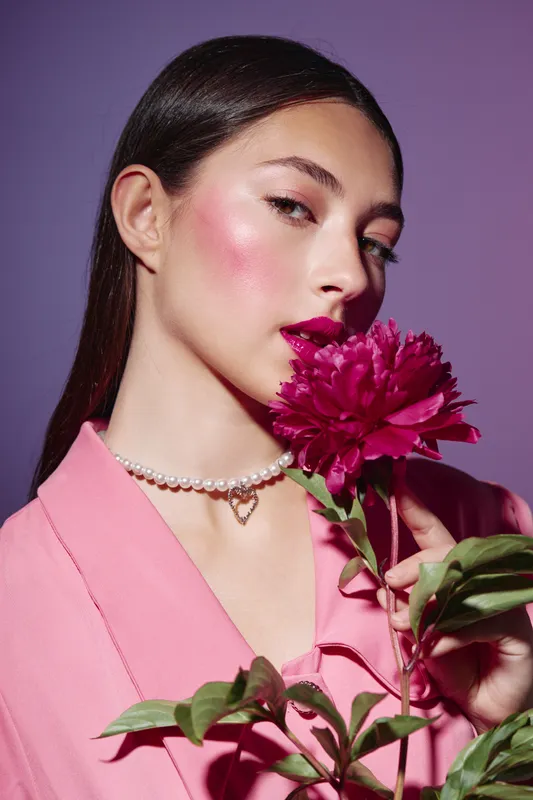 Fashionable young woman wearing pink blazer with bright makeup poses holding a fuchsia flower. She wears a pearl necklace against a purple background. Beauty and creative fashion portrait.