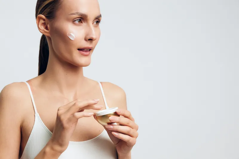 Woman skincare face cream applying with fingers, young woman holds moisturizer jar, calm beauty routine, studio white background, soft natural light, hygiene and wellness care, facial beauty concept