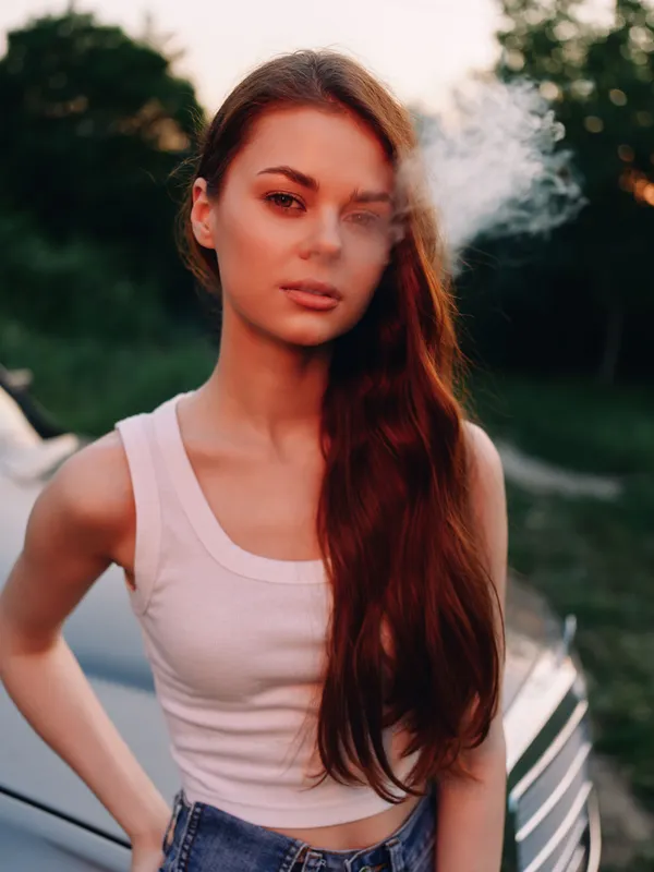 Young woman smoking with a sultry expression, wearing a white tank top, in an outdoor setting at sunset, with a blurred car in the background.