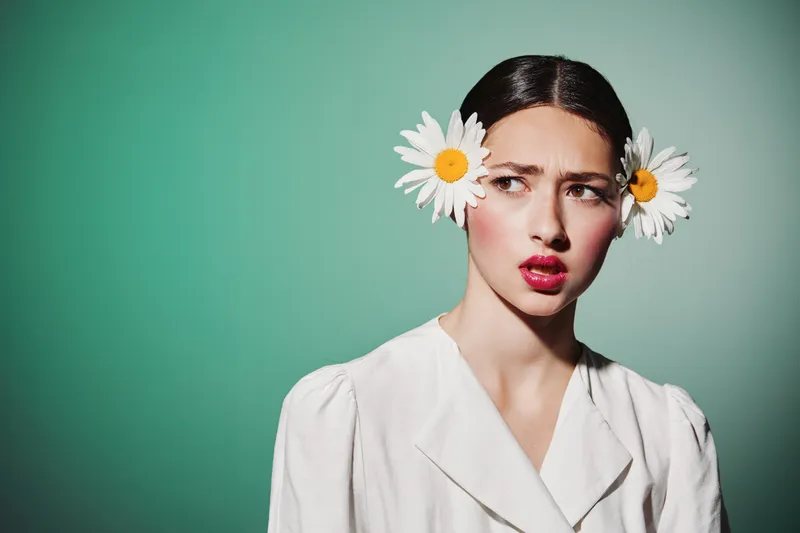 Young woman in white posing with bright makeup and natural daisies in hair against pastel green background. Concept of beauty, creative fashion, floral style, and vibrant portrait.