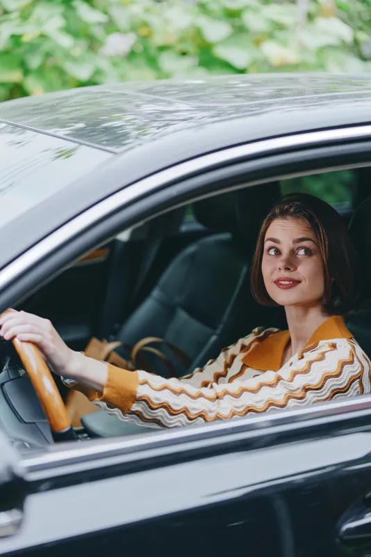 A stylish woman sits behind the wheel of a dark car, smiling softly as she gazes ahead, capturing confidence and chic mood.