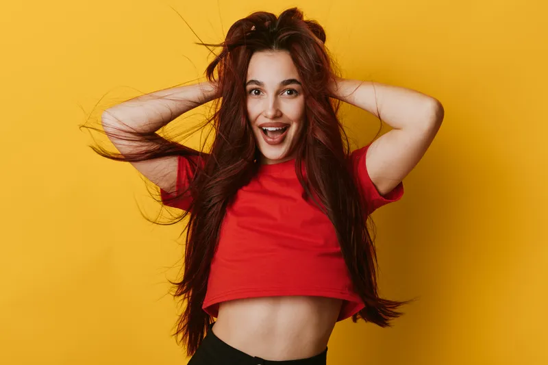 Young woman with long hair wearing red crop top posing with hands in hair and smiling against yellow background for lifestyle, happiness and fashion portrait.
