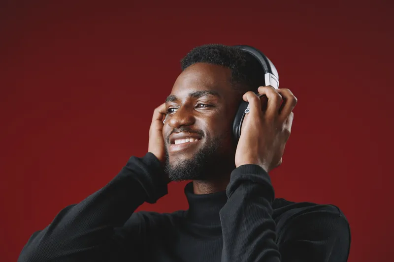 Young African man enjoying music with headphones against a red background, expressing happiness and relaxation, ideal for wellness and leisure themes