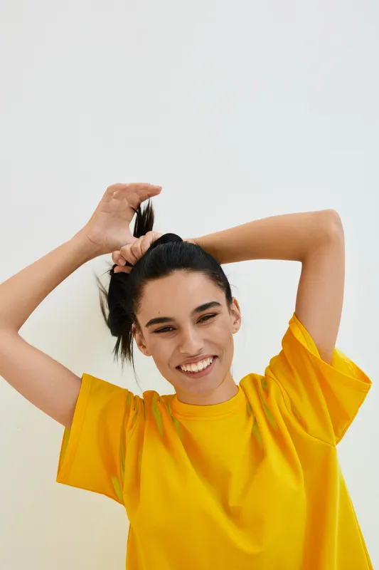smiling woman in bright yellow t-shirt joyfully tying hair up against plain background, happy young adult expressing positivity and energy