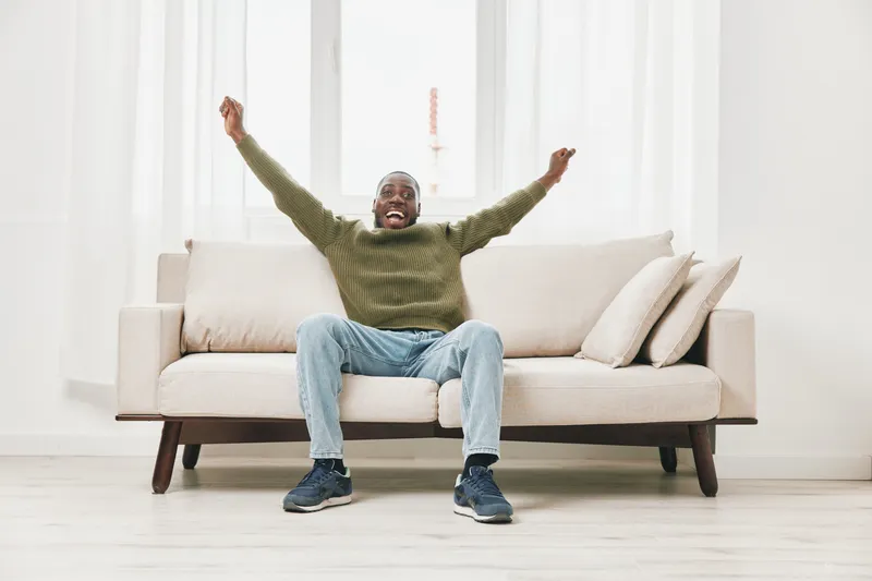Happy African American man celebrating at home on a cozy sofa with casual outfit and bright interior, expressing joy and relaxation in his living space