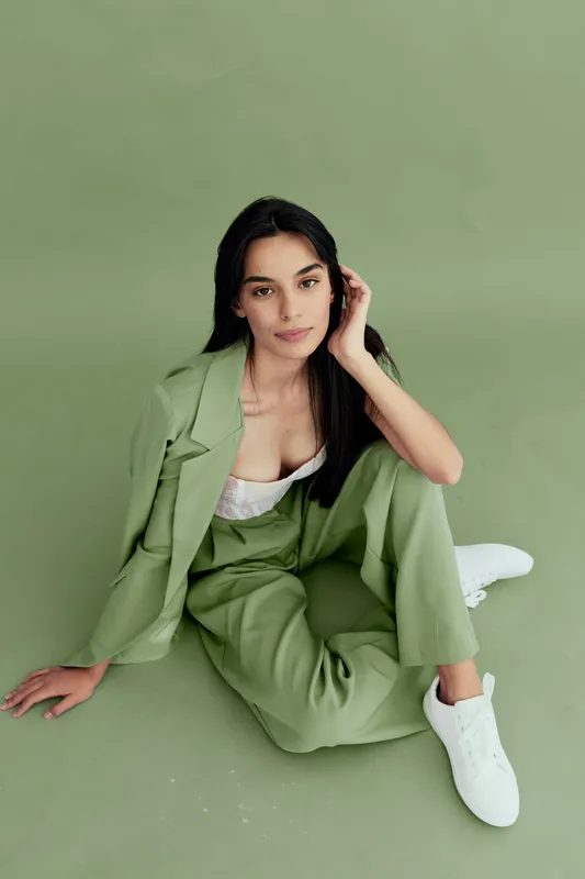 Young woman sitting on floor in stylish light green suit with white sneakers, posing indoors against a matching green background in a modern minimalist fashion setup.