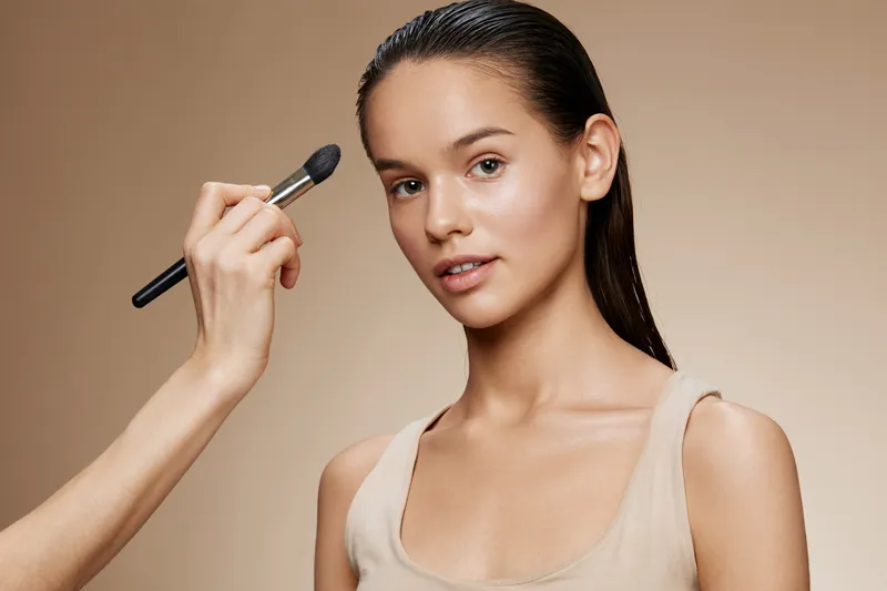 Portrait of a beautiful young model receiving makeup application with a neutral background, showcasing natural beauty and professionalism in cosmetics