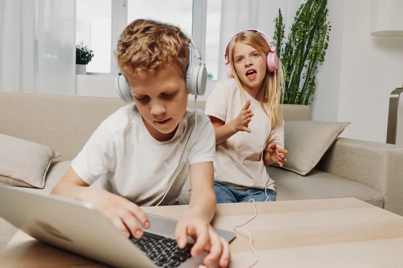 Two children wearing headphones interact with laptop in living room, boy focused on device while girl sings and gestures behind him.