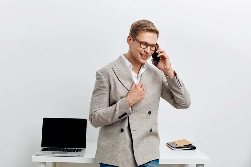 Smiling man in glasses talking on phone while standing at desk in modern office interior. Casual business attire, confident expression, technology gadgets, office supplies, bright workspace, professio
