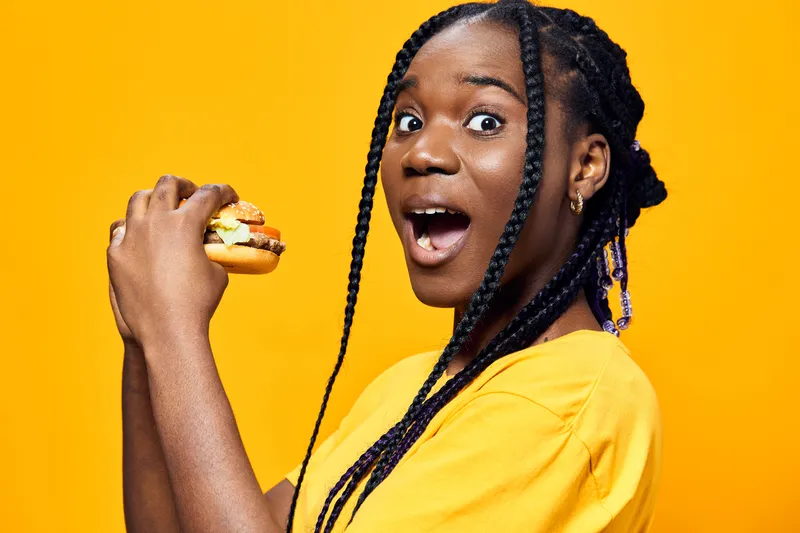 Excited young woman of African descent holding a delicious burger, showcasing joy against a bright yellow background Perfect for food and lifestyle themes