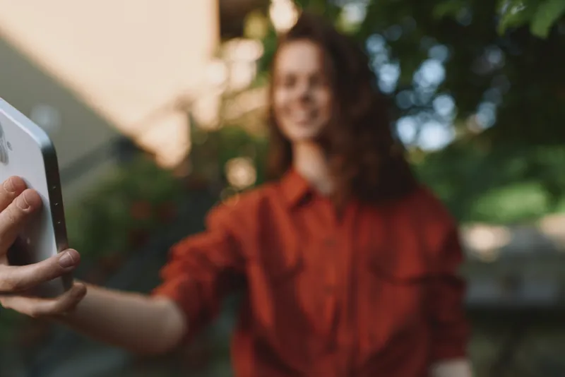 Young woman in a red shirt takes a casual outdoor selfie, smiling softly as warm light and green background frame the moment