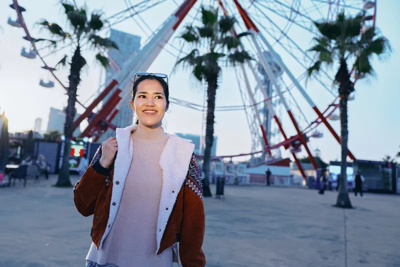 Woman in a cozy sweater and jacket stands in a park with a ferris wheel in the background, casual fashion, urban vibe, warm evening light, candid outdoor scene
