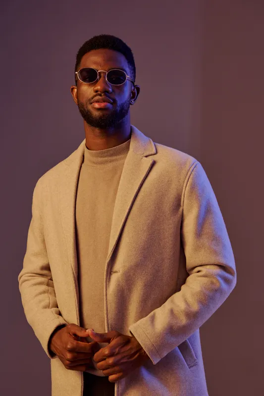 Stylish young man wearing a beige coat and sunglasses, showcasing confidence and modern fashion in a softly lit studio setting