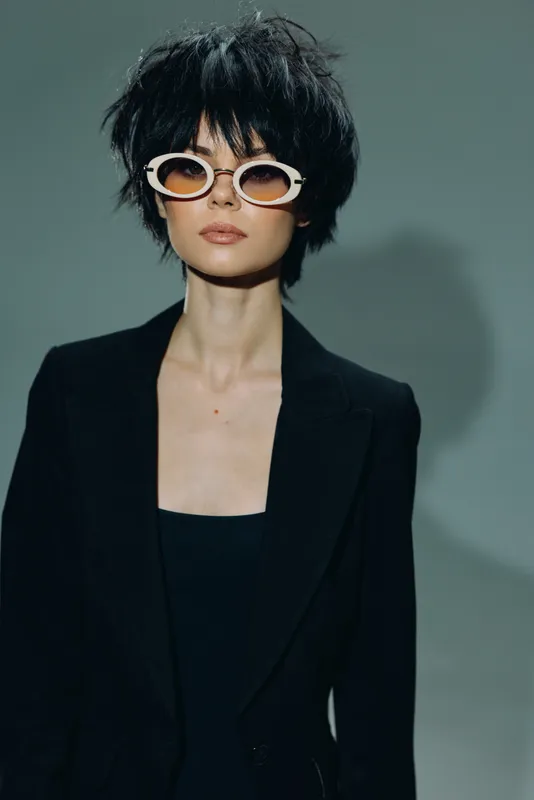 Stylish woman with short black hair and unique white sunglasses wearing a black blazer against gray background
