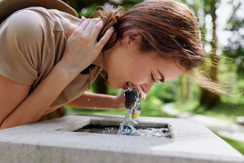Woman drinking water from a public fountain in a sunny park, young adult closeup in casual shirt leaning to sip from stone tap, hydration, refreshment and leisure.